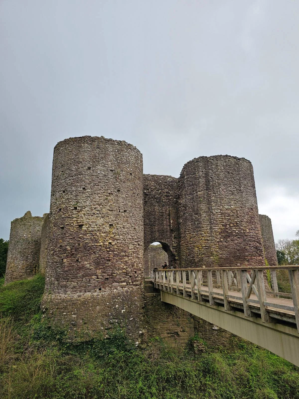 Large stone castle with two round towers and an arched entrance, connected to a wooden bridge over a moat, under a cloudy sky.