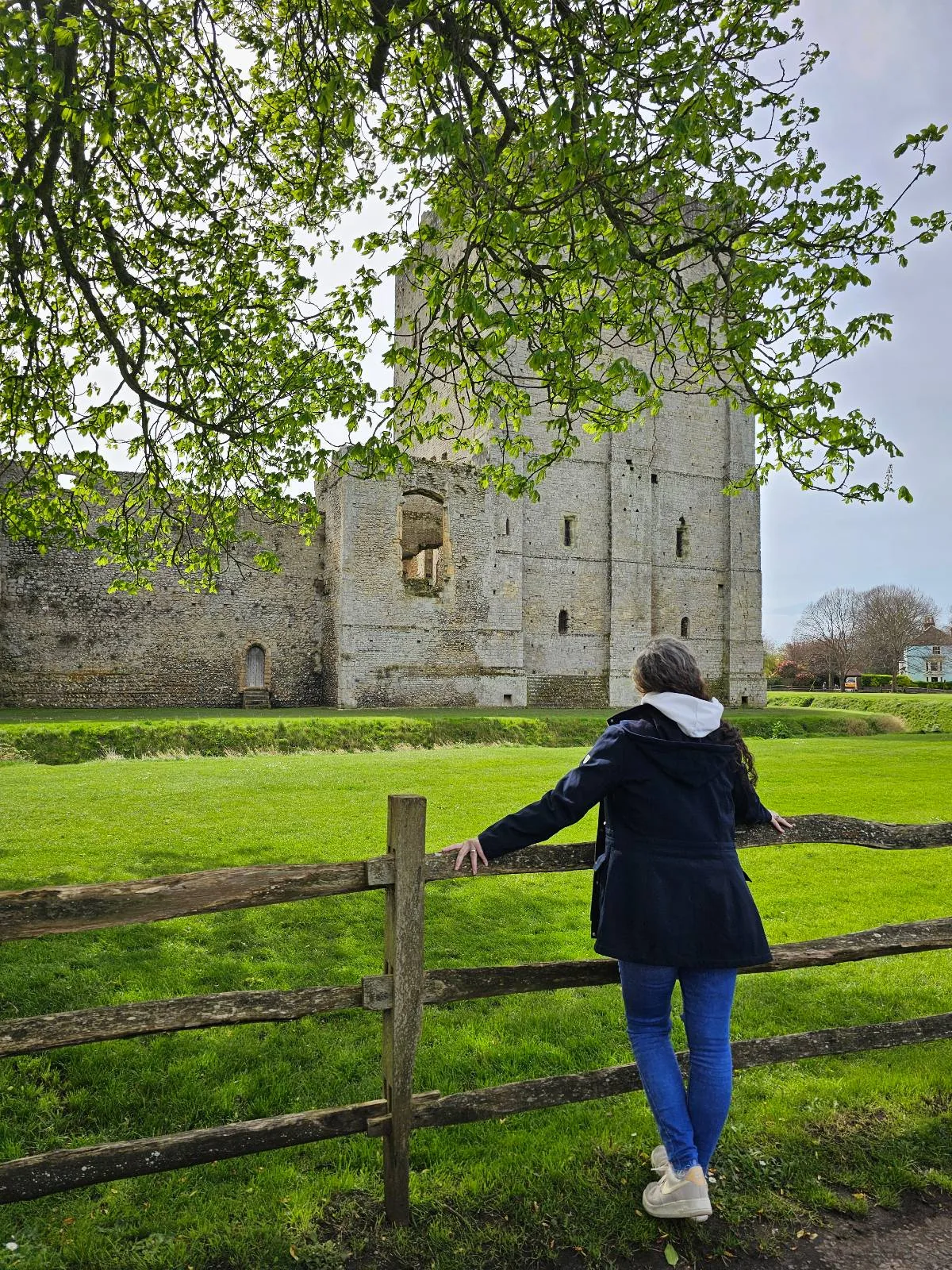 A person in a dark coat and blue jeans leans on a wooden fence, looking at an old stone building surrounded by greenery, with tree branches overhead.