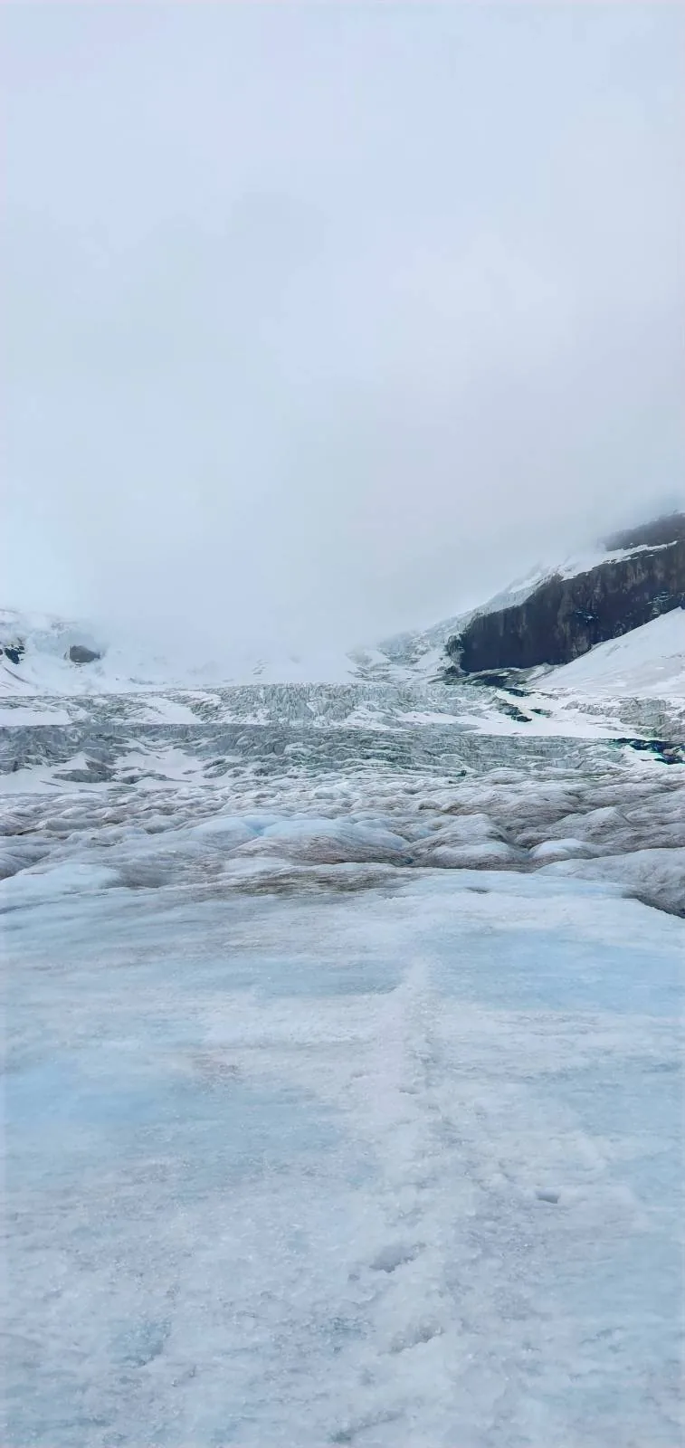 A snowy, icy landscape with a glacier stretching into the distance under a cloudy sky, flanked by dark rocky hills.