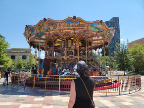 A person observing a traditional carousel in an urban outdoor setting on a sunny day.