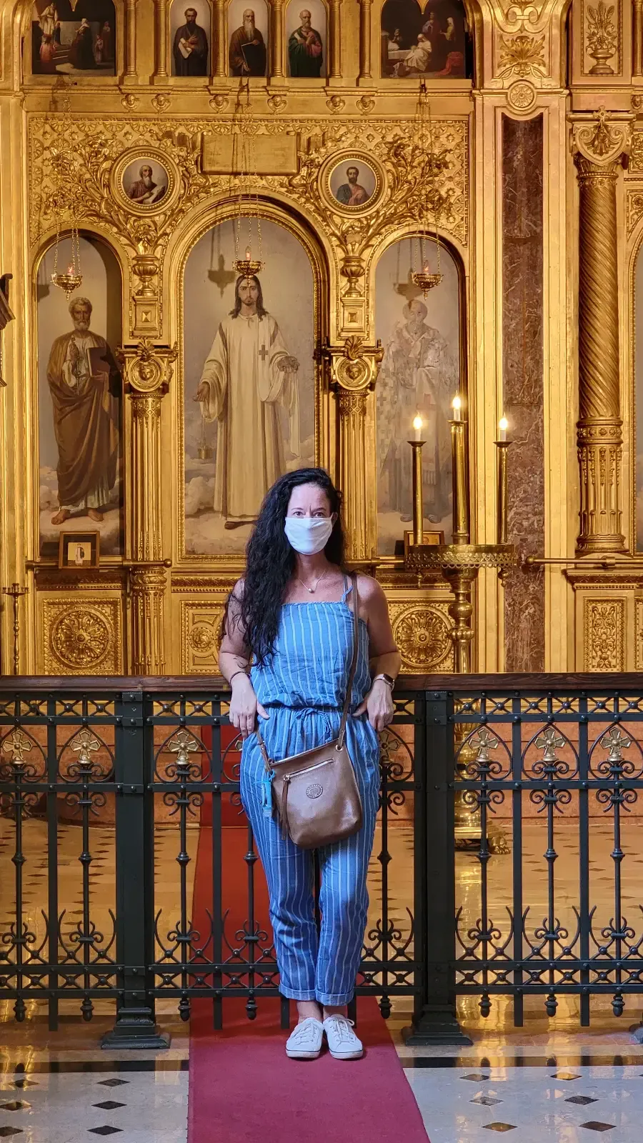 A woman with long dark hair stands on a red carpet in front of an ornate, golden religious altar, wearing a blue outfit and a face mask, with a brown crossbody bag.