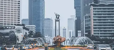 A wide city street in Jakarta featuring the Selamat Datang Monument at a large roundabout with fountains, surrounded by modern glass skyscrapers under an overcast sky.