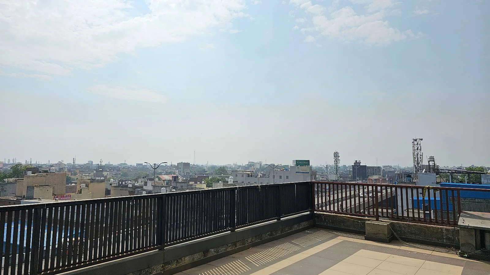 A rooftop terrace with a black railing overlooks a cityscape under a partly cloudy sky. Buildings stretch into the distance, and the weather appears clear and sunny.