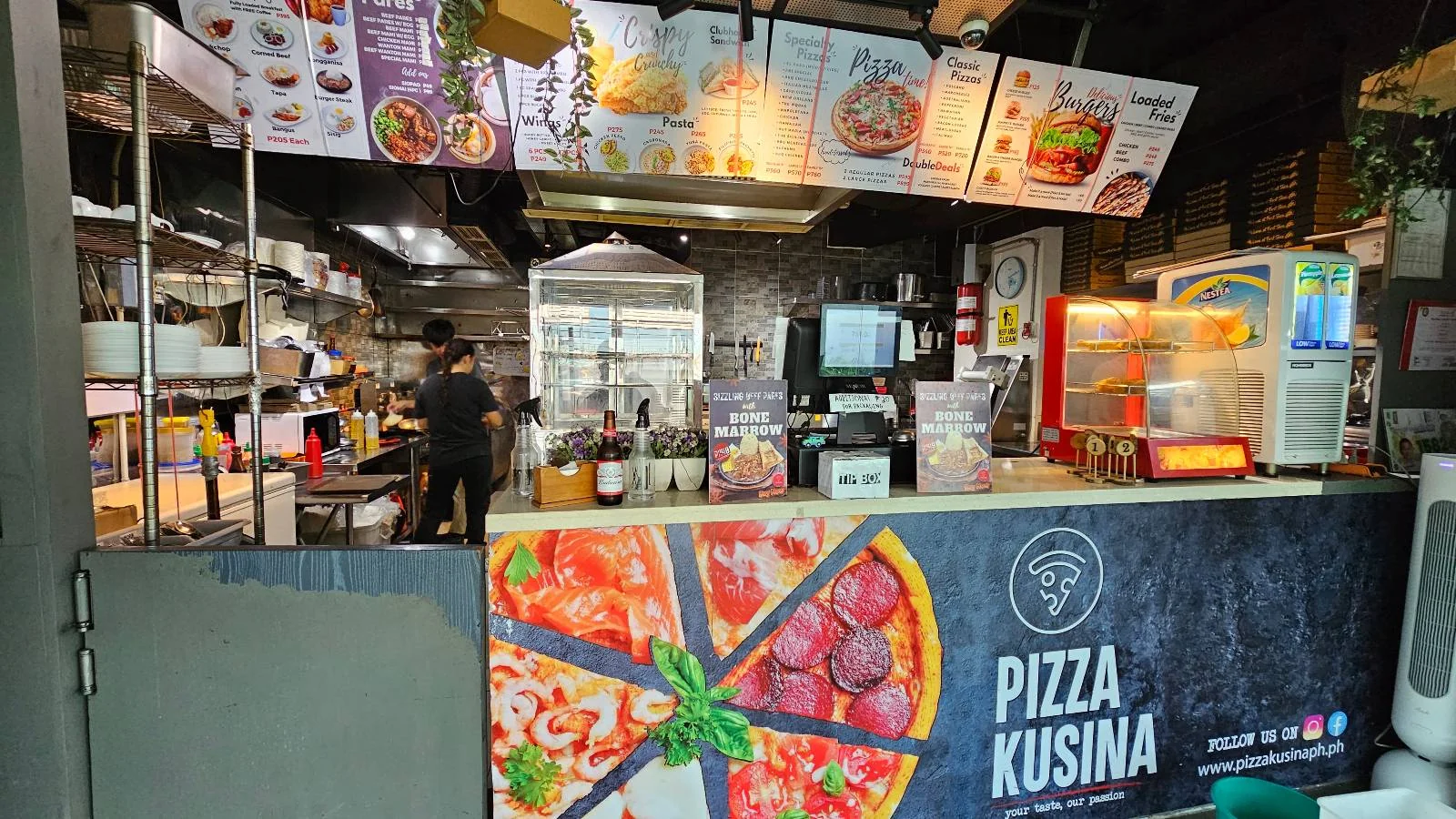 A food stall with a large "Pizza Kusina" sign, menu boards above the counter, a pizza graphic, and kitchen equipment visible in the background. One person is working behind the counter.