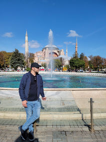 A man in casual attire stands by a fountain, with the architectural brilliance of the Hagia Sophia and its minarets in the background under a clear blue sky.