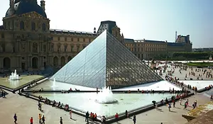 The glass Louvre Pyramid in Paris surrounded by historic palace buildings with a water fountain in front under a bright sky.