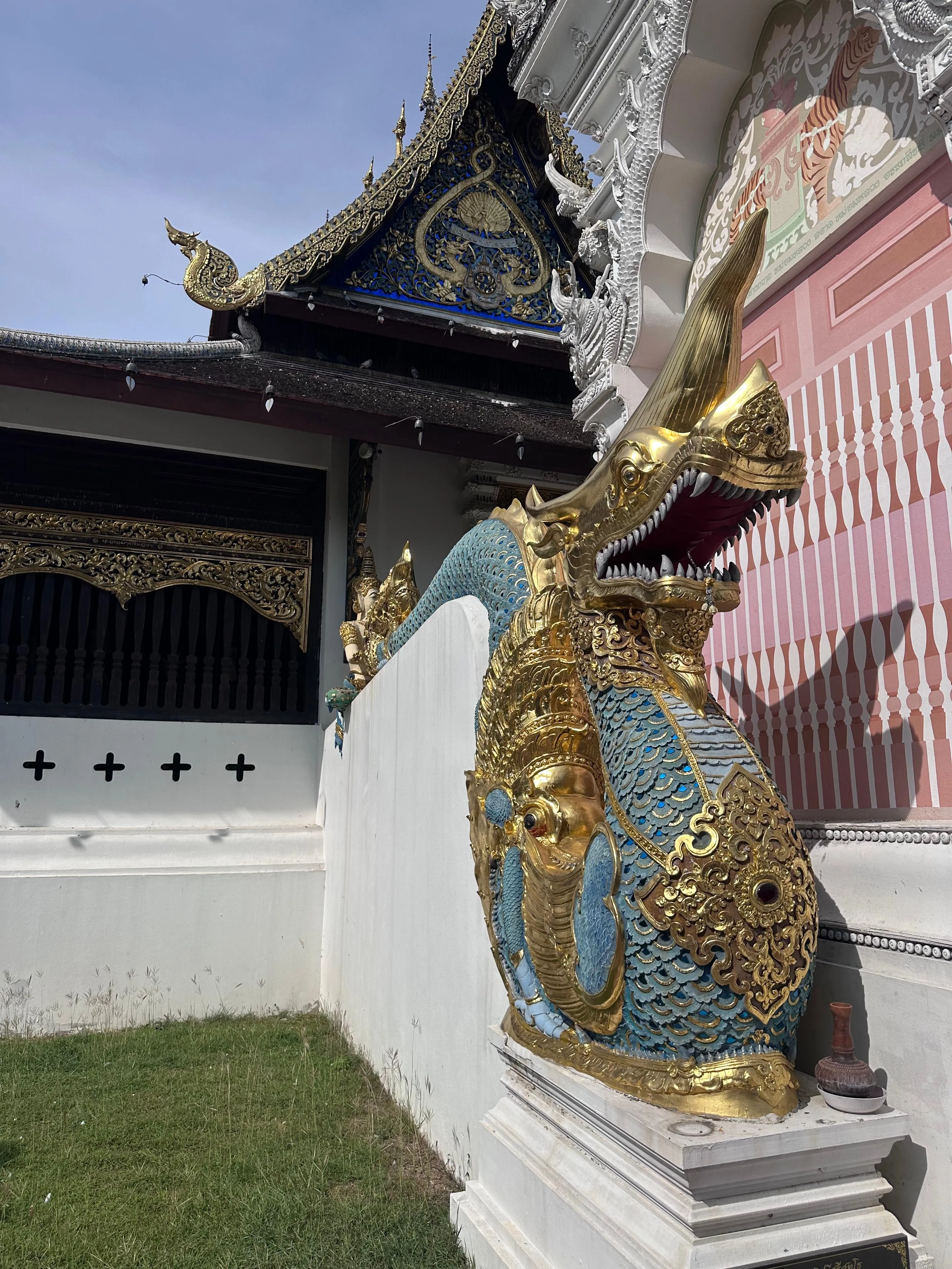 Ornate dragon sculpture with gold, blue, and green detailing decorates the white exterior of a traditional Thai temple under a blue sky. Pink and black walls are visible in the background.