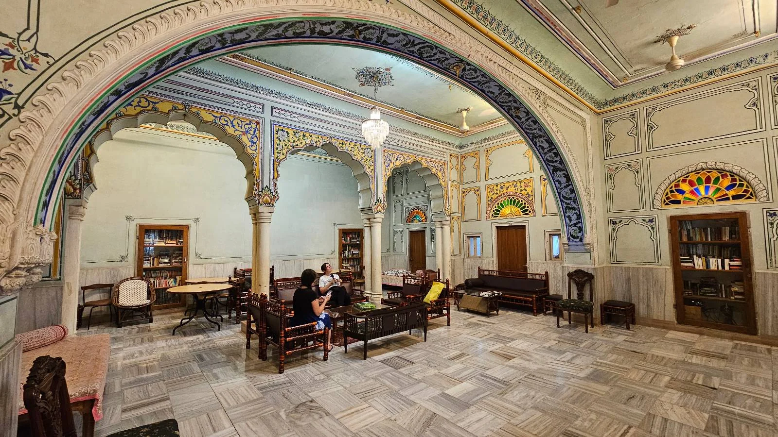 A spacious room with ornate arches, colorful stained glass windows, and high ceilings. Several people sit on wooden chairs and sofas, reading or conversing. The décor is elegant and traditional.