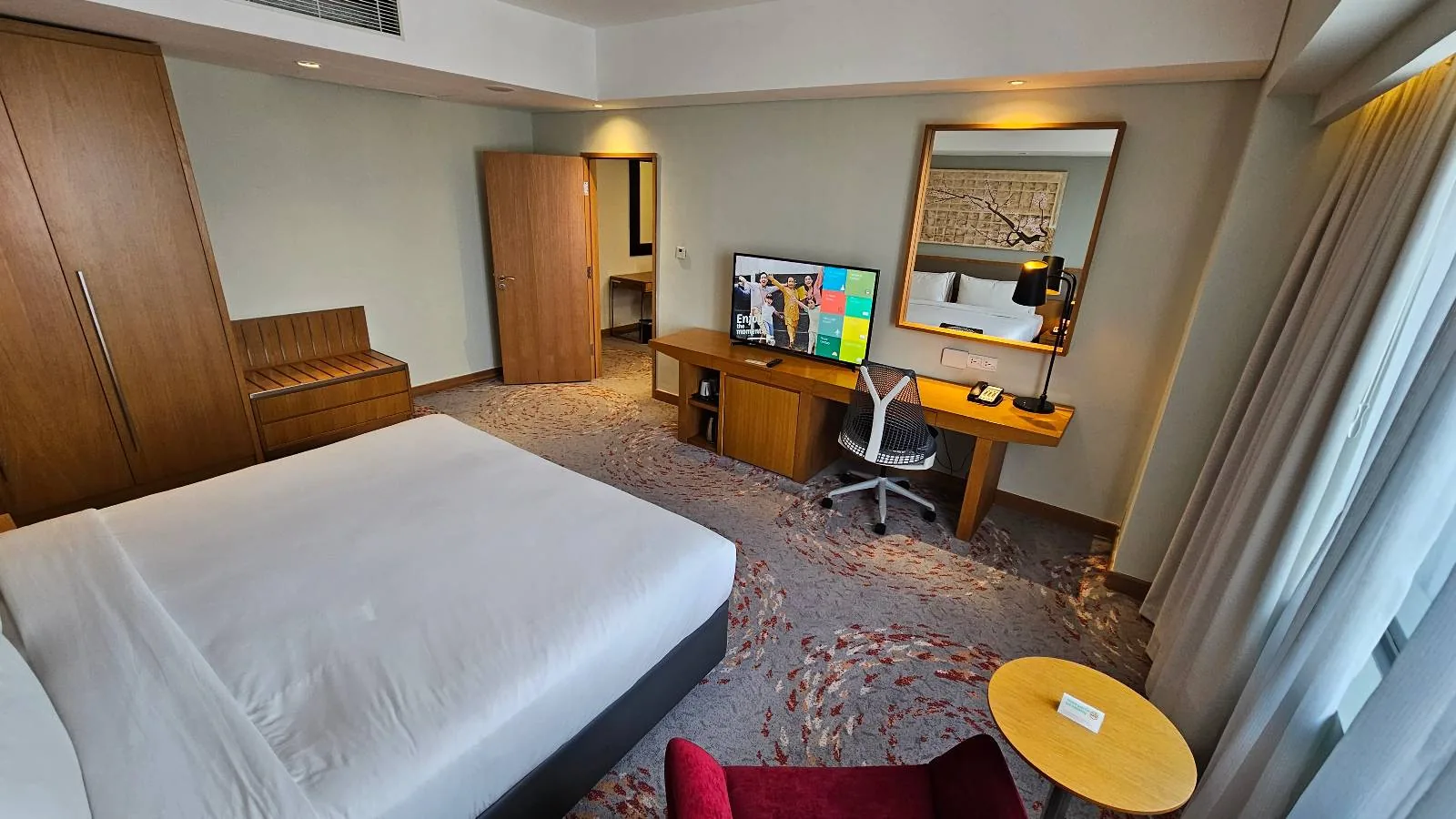A modern hotel room with a large bed, a TV on a wooden desk, a white office chair, a wardrobe, a full-length mirror, a small round table, and a red armchair near a window with curtains.