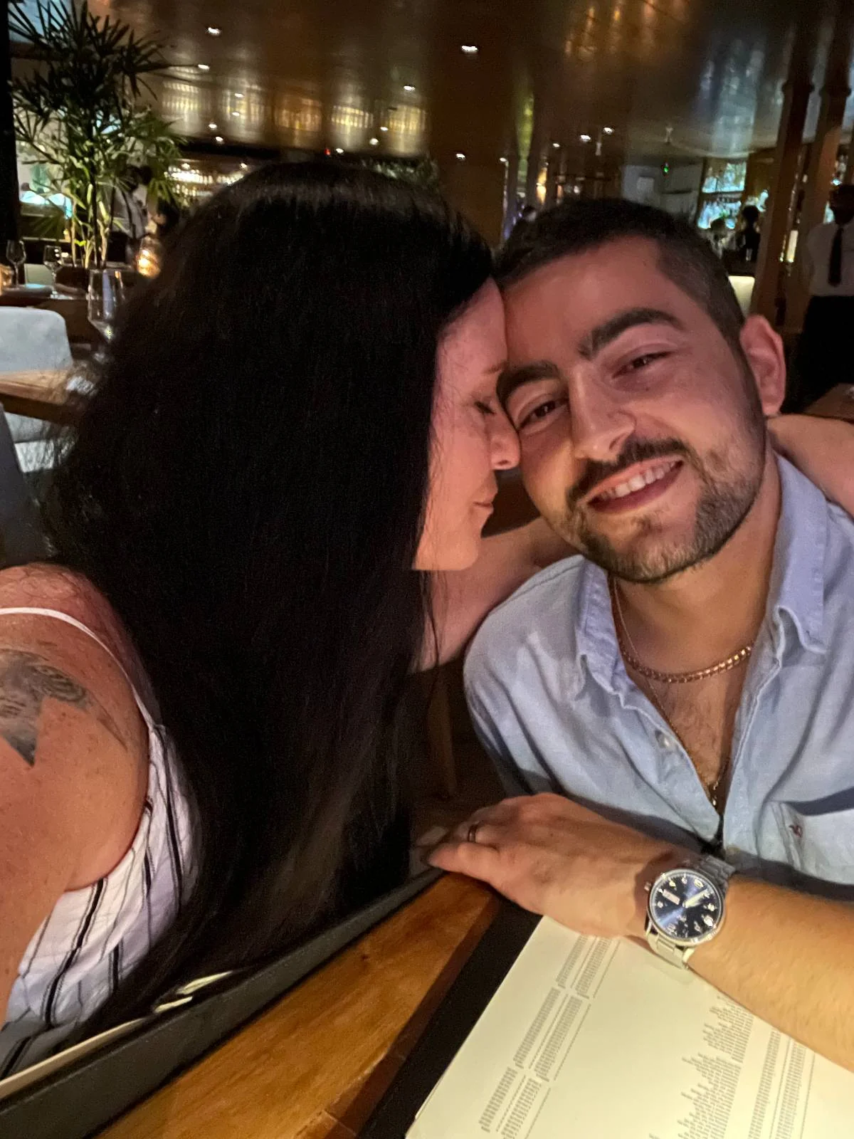 A woman with long dark hair leans affectionately towards a smiling man with short hair and a beard, both sitting at a restaurant table with menus visible. The setting is warmly lit, creating a cozy atmosphere.