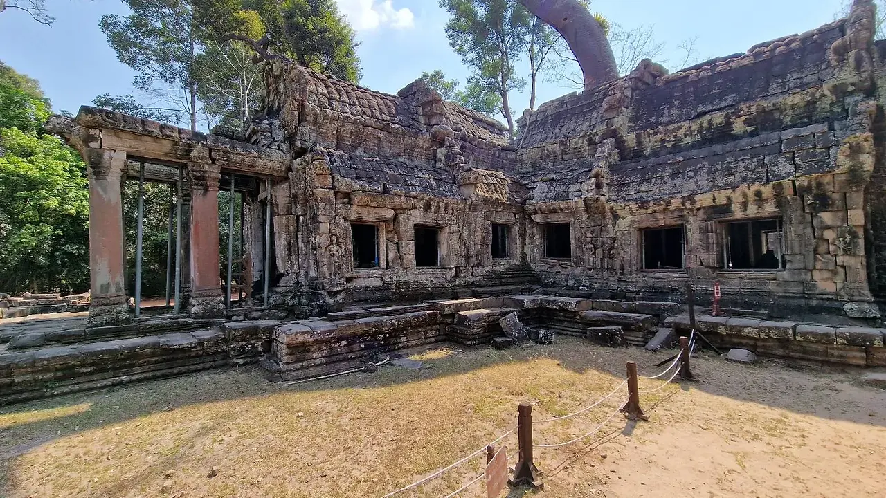 Wide-angle view of the ancient stone ruins of Ta Prohm temple in Cambodia, featuring a dirt courtyard, weathered stone structures, a wooden viewing platform with steps, and towering jungle trees.