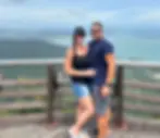 Couple posing on a scenic viewpoint with ocean and hills in the background. They are smiling, wearing casual summer outfits and sunglasses.
