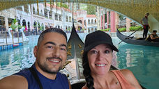 A smiling man and woman take a selfie while sitting in a gondola on a blue canal inside a shopping mall with shops and a gondolier in the background under a decorative bridge.