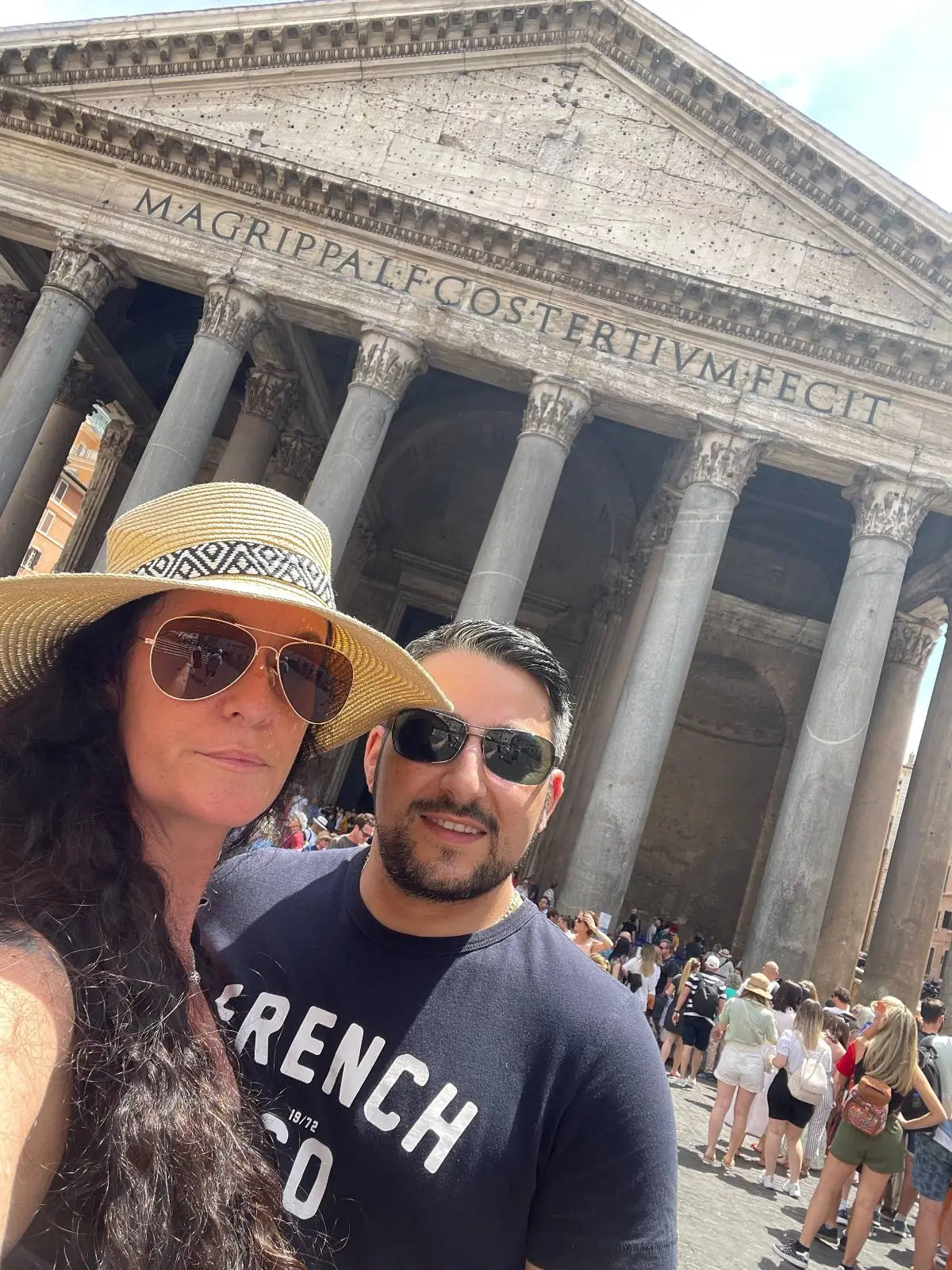 A woman in a sunhat and sunglasses and a man in sunglasses pose for a selfie in front of the ancient Pantheon with large columns, surrounded by a crowd of tourists.