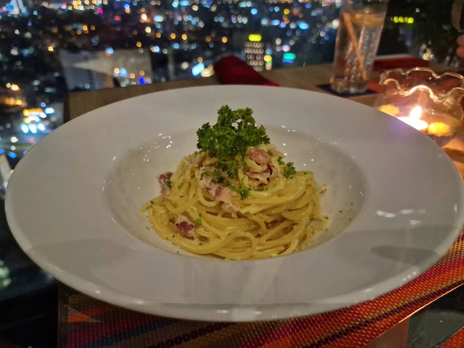 A plate of creamy pasta garnished with parsley sits on a table, with a cityscape full of colorful lights blurred in the background and a lit candle nearby.