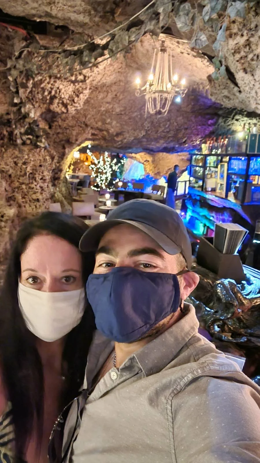 A man and woman wearing masks take a selfie in a cavern-themed restaurant. The background features textured rock walls, a chandelier, and lighted decorations, creating a cozy atmosphere.