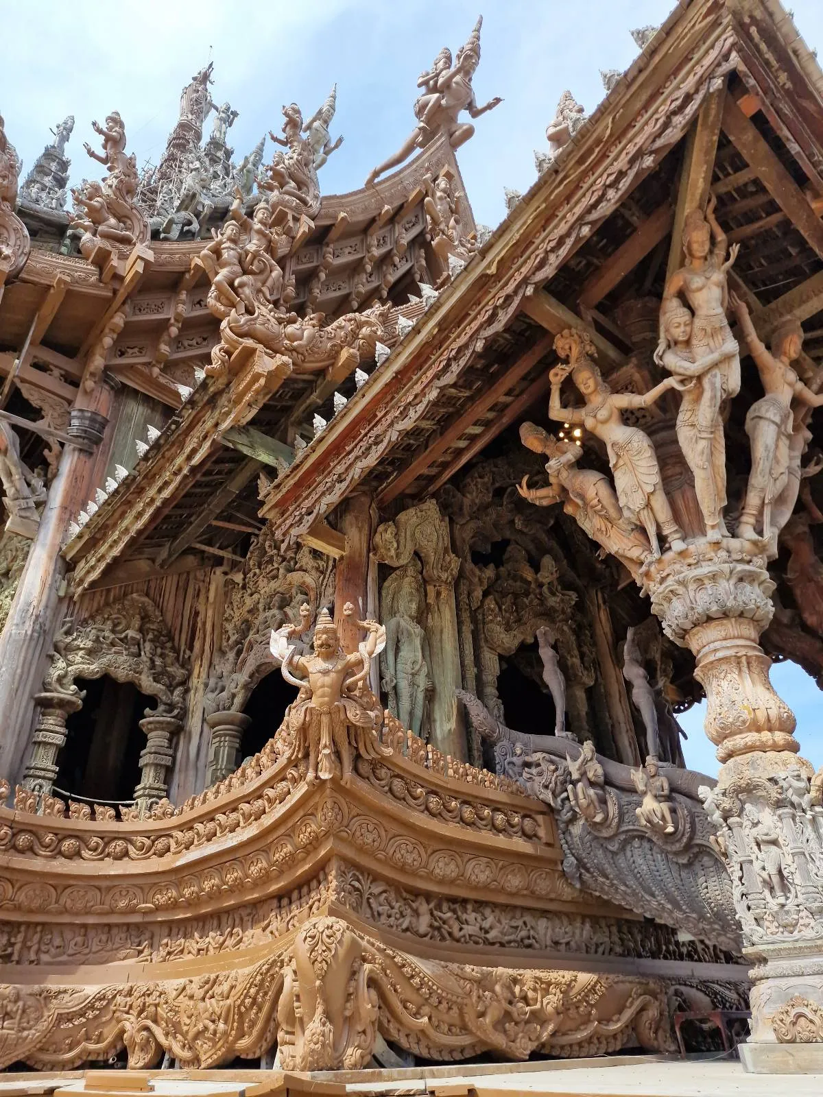 Ornate wooden temple with intricate carvings of figures and patterns, featuring detailed columns and roof decorations against a blue sky.