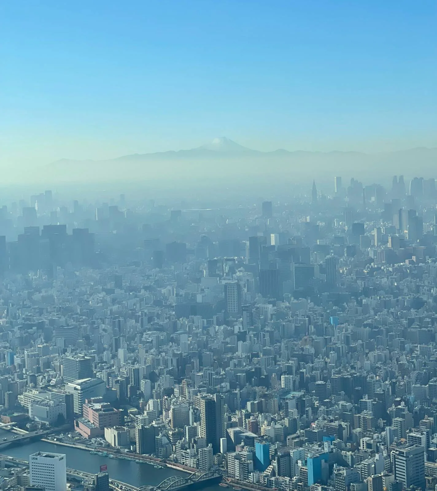 Aerial view of a sprawling cityscape with a clear blue sky. In the distance, a mountain peak emerges through a layer of haze, adding depth to the urban scene. The buildings vary in height and density across the city.