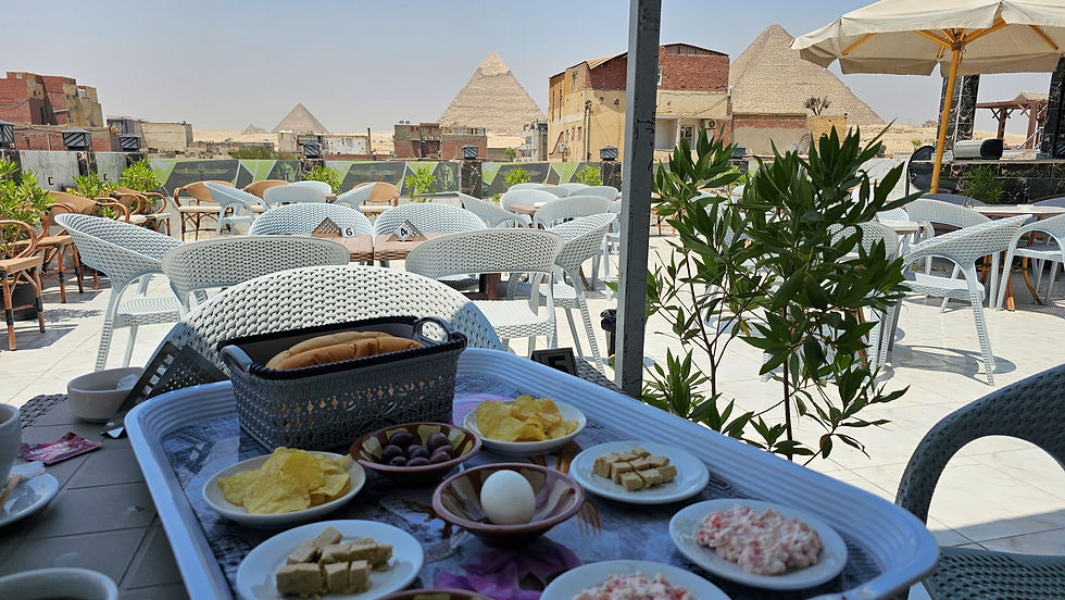 Outdoor cafe table with breakfast items, chairs, and greenery. Pyramids visible in the background under a clear blue sky.