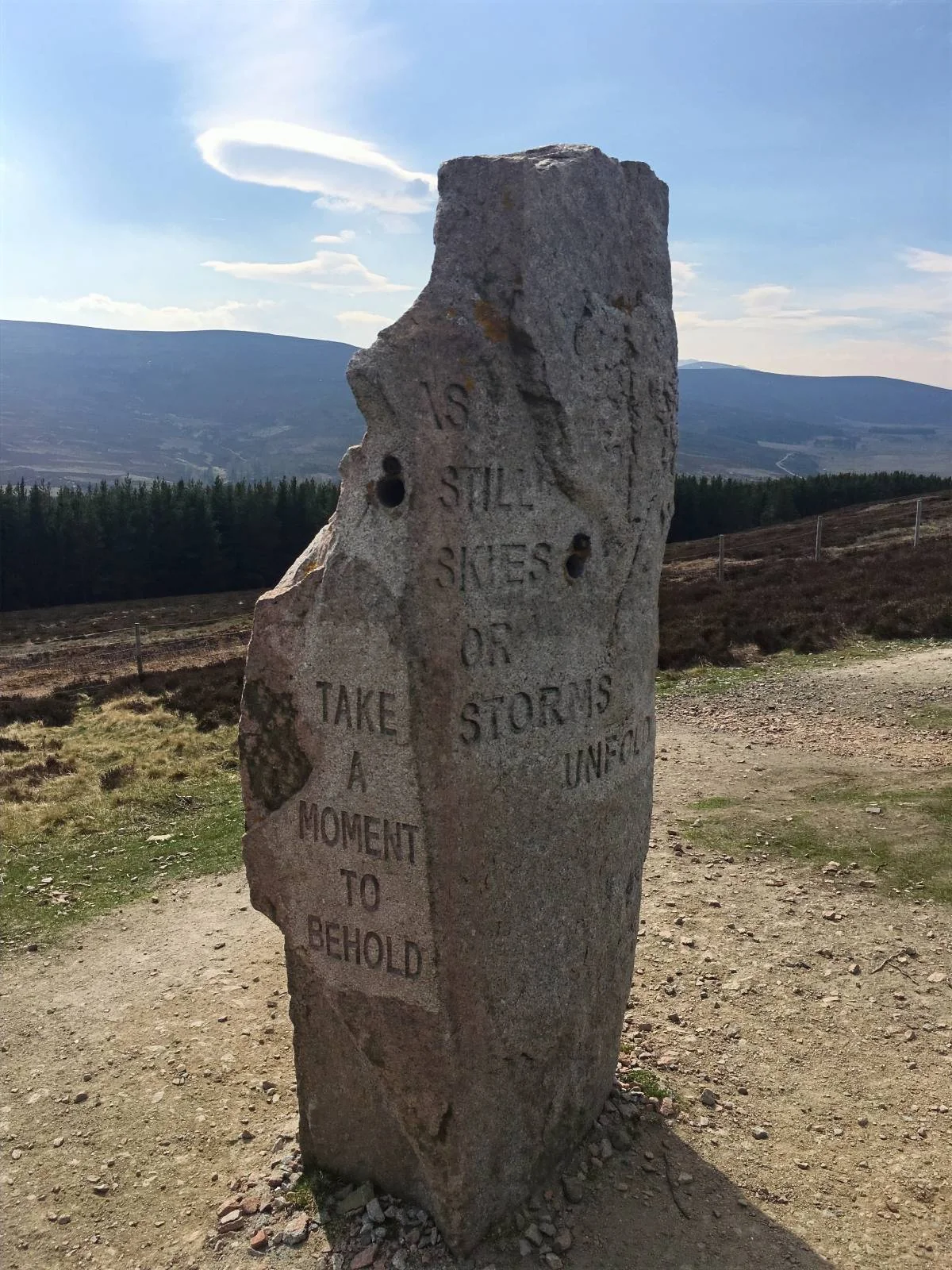 A stone marker with the text "Take a moment to behold" stands on a dirt path in a scenic mountainous area under a blue sky. In the background, metal sculptures can be seen.