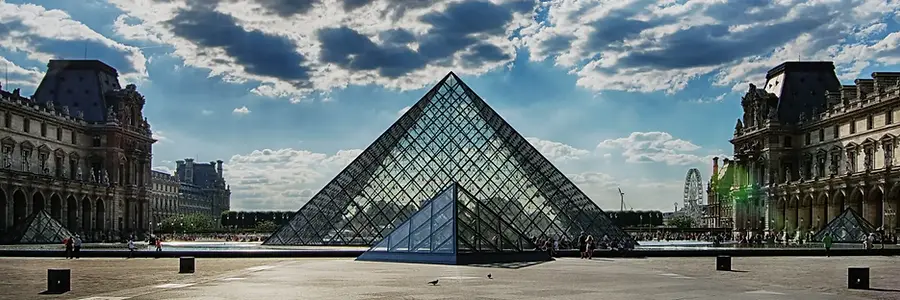 The glass Louvre Pyramid in Paris surrounded by historic palace buildings under a partly cloudy sky.