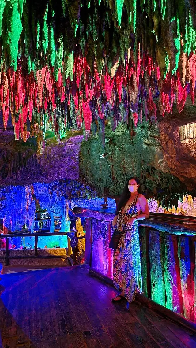 A person stands on a wooden platform inside a colorful cave decorated with hanging, illuminated stalactites in shades of green, pink, and purple. They are wearing a long dress and holding a small bag. The atmosphere is vibrant and mystical.