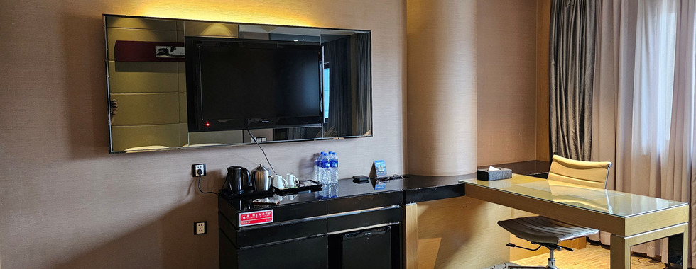 A modern hotel room features a wall-mounted flat-screen TV, a desk with a chair, a minibar with drinks, and large windows with curtains letting in natural light.
