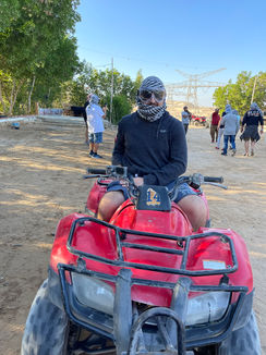 A person wearing sunglasses and a patterned headscarf sits on a red ATV, perhaps enjoying one of the thrilling ATV tours. They are outdoors on a dirt path with trees and power lines in the background, reminiscent of El Gouna's adventurous trails. Other people are walking in the distance.