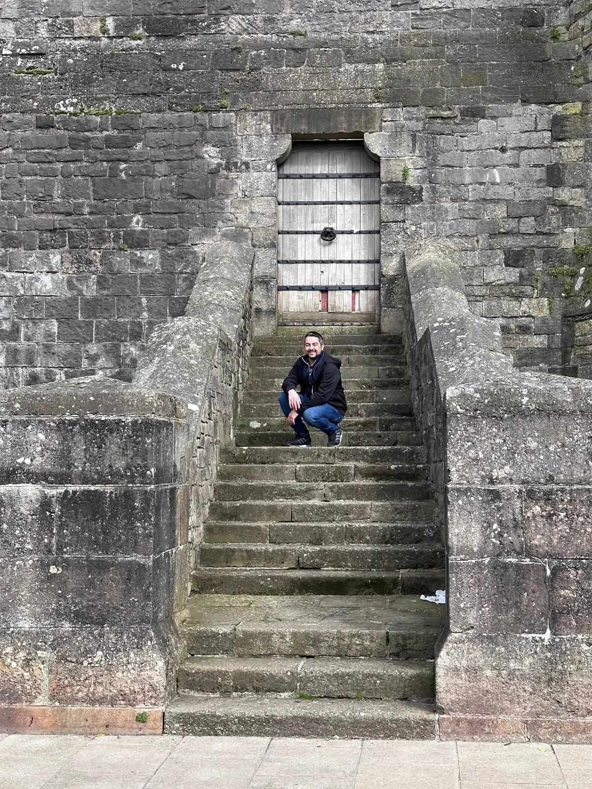 A person crouches on stone steps leading to a closed metal door set in an old, weathered stone wall.