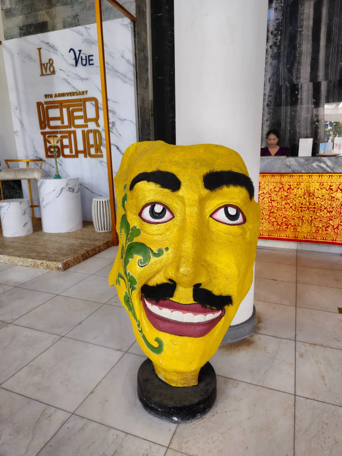 A large, vibrant yellow sculpture of a human head with exaggerated facial features, including a mustache and a decorative green swirl by the eye, is displayed in a lobby. A reception desk is visible in the background.