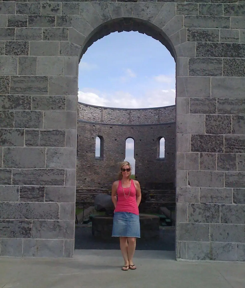 Woman in pink top and denim skirt stands smiling under stone archway, with a circular stone ruin and blue sky in the background.