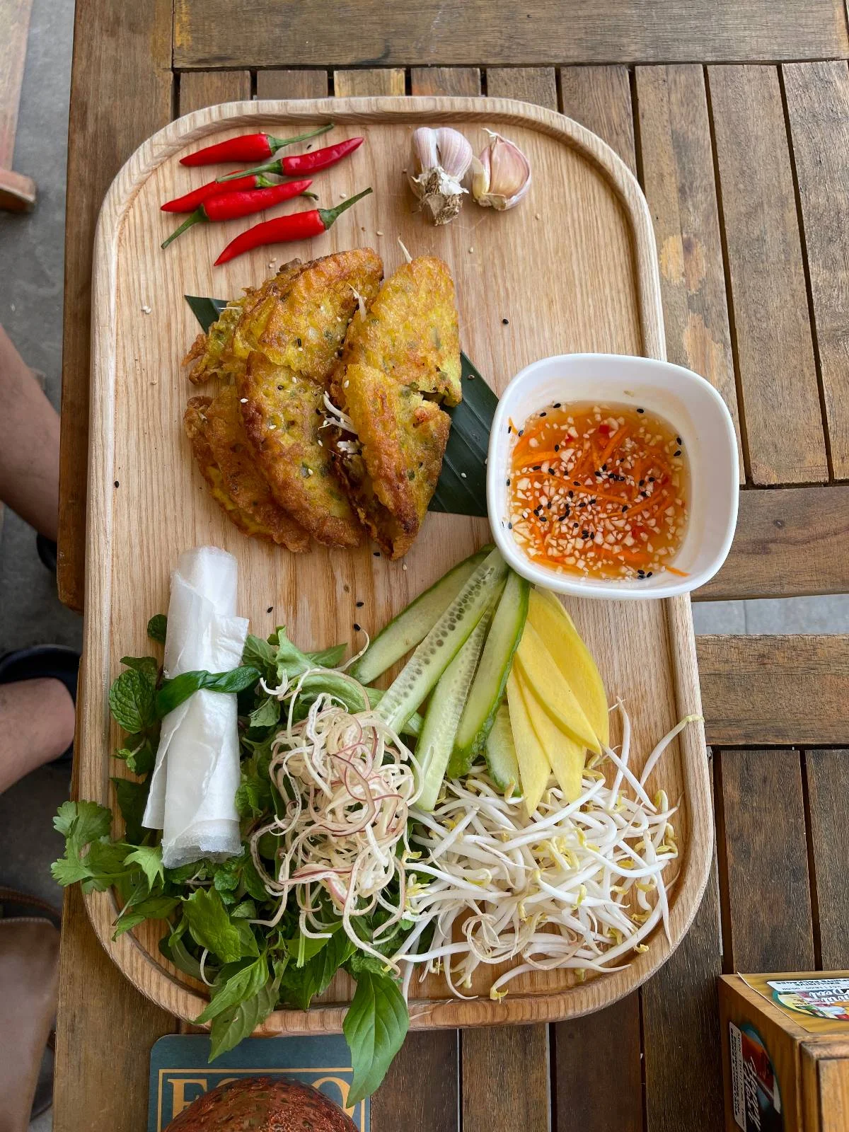 A wooden tray with Vietnamese pancakes, a bowl of dipping sauce, fresh red chili peppers, garlic cloves, herbs, cucumber slices, mango slices, and bean sprouts. Wrapped rice paper is placed on a leaf.