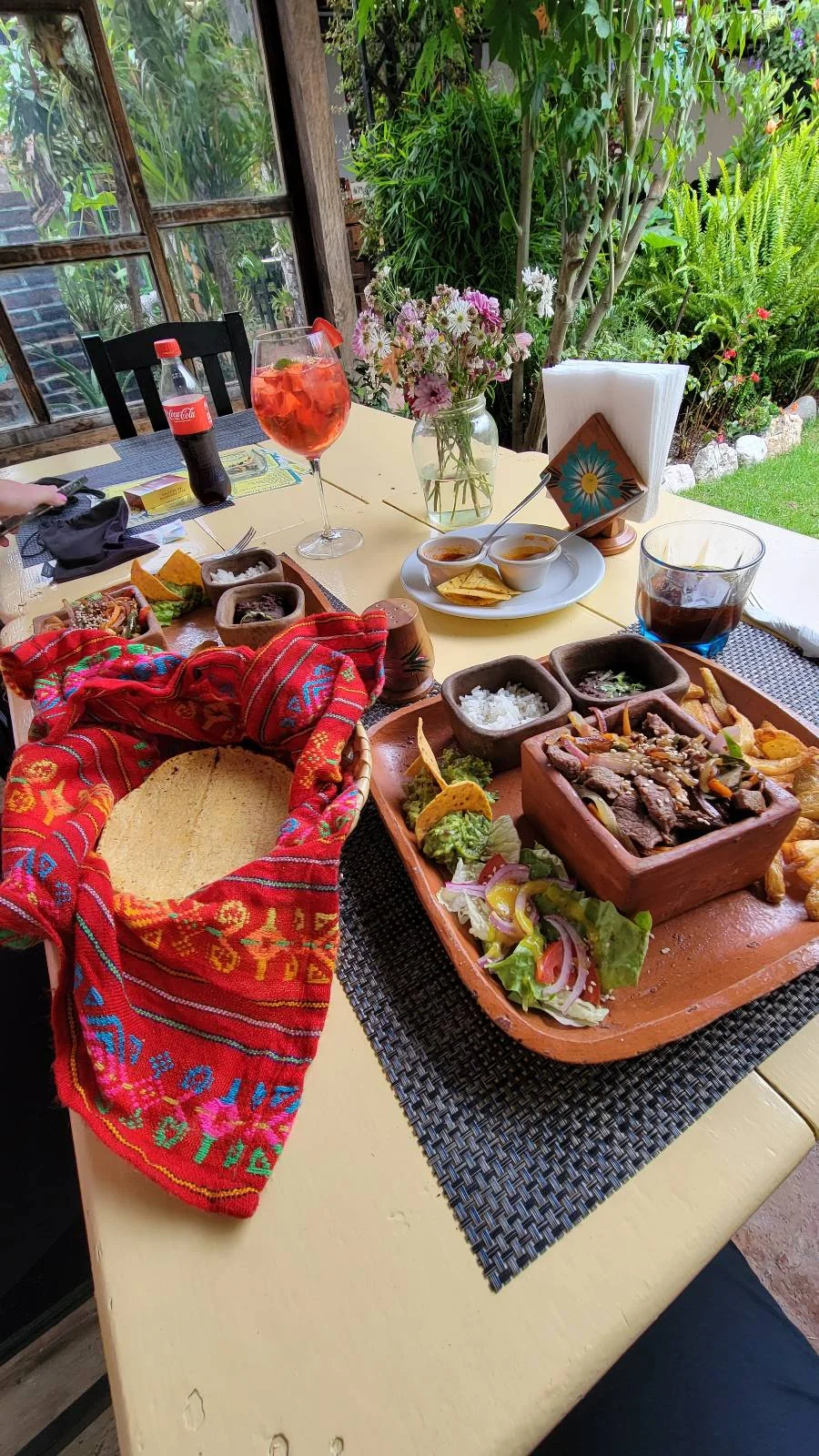 A table set outdoors with a meal. There's a platter of fajitas with grilled vegetables, beans, and sauces. Next to it is a basket covered with a colorful cloth. Drinks include cola and a cocktail. Flowers decorate the corner of the table.