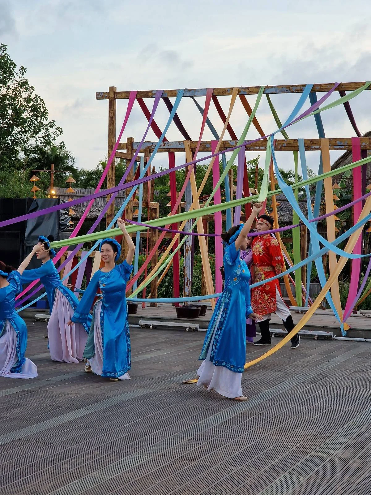Women in traditional blue and red attire performing a dance on an outdoor stage with a colorful geometric backdrop.