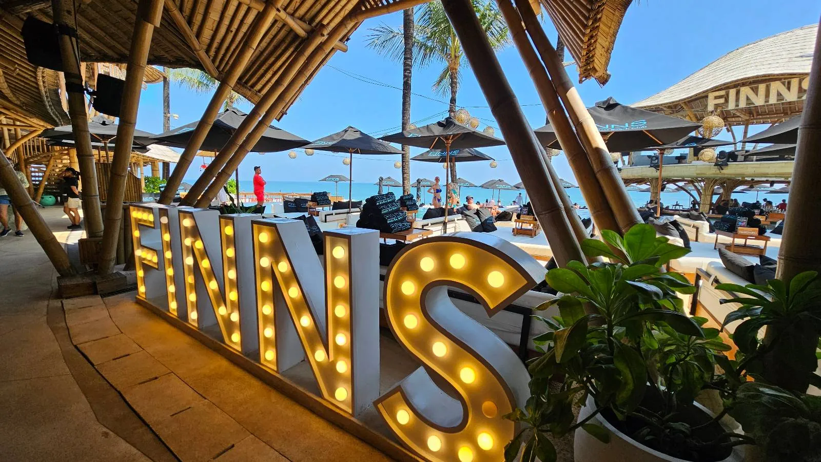 Large illuminated "FINNS" sign at a bamboo beach club. People relax on loungers under umbrellas. Ocean view with clear blue sky.