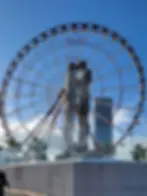 Two metallic sculptures intertwine in front of a large Ferris wheel under a clear blue sky. Palm trees and a tall building are visible.