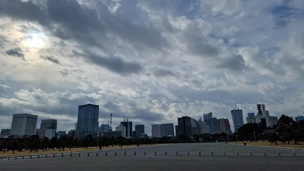Cloudy sky over city skyline, skyscrapers in the distance. Dark foreground with scattered trees. Sun peeks through clouds, creating a moody scene.