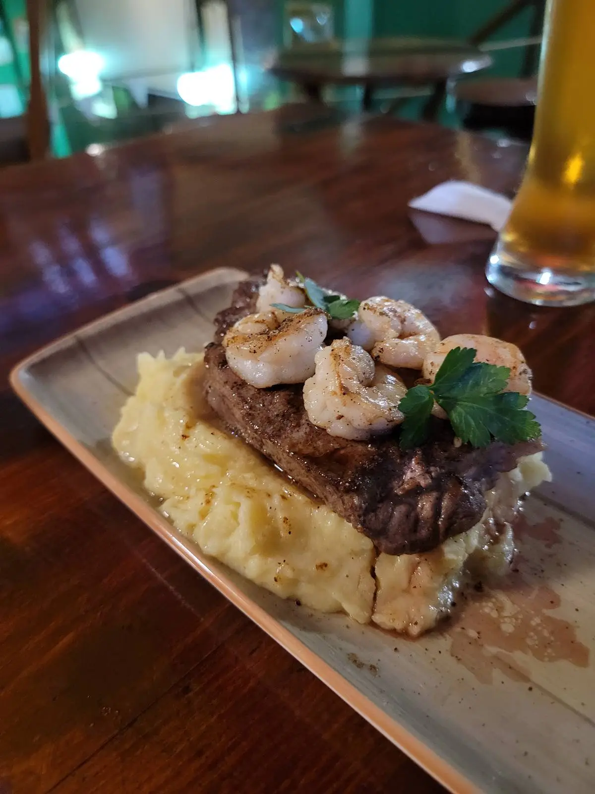 A rectangular plate with a steak topped with shrimp and herbs, served on a bed of mashed potatoes. A glass of beer is in the background on a wooden table.