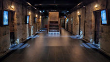 Dimly lit Bodmin Jail cell block with historical exhibit posters on stone walls and a central staircase leading upwards, marked by an "exit this way" sign.