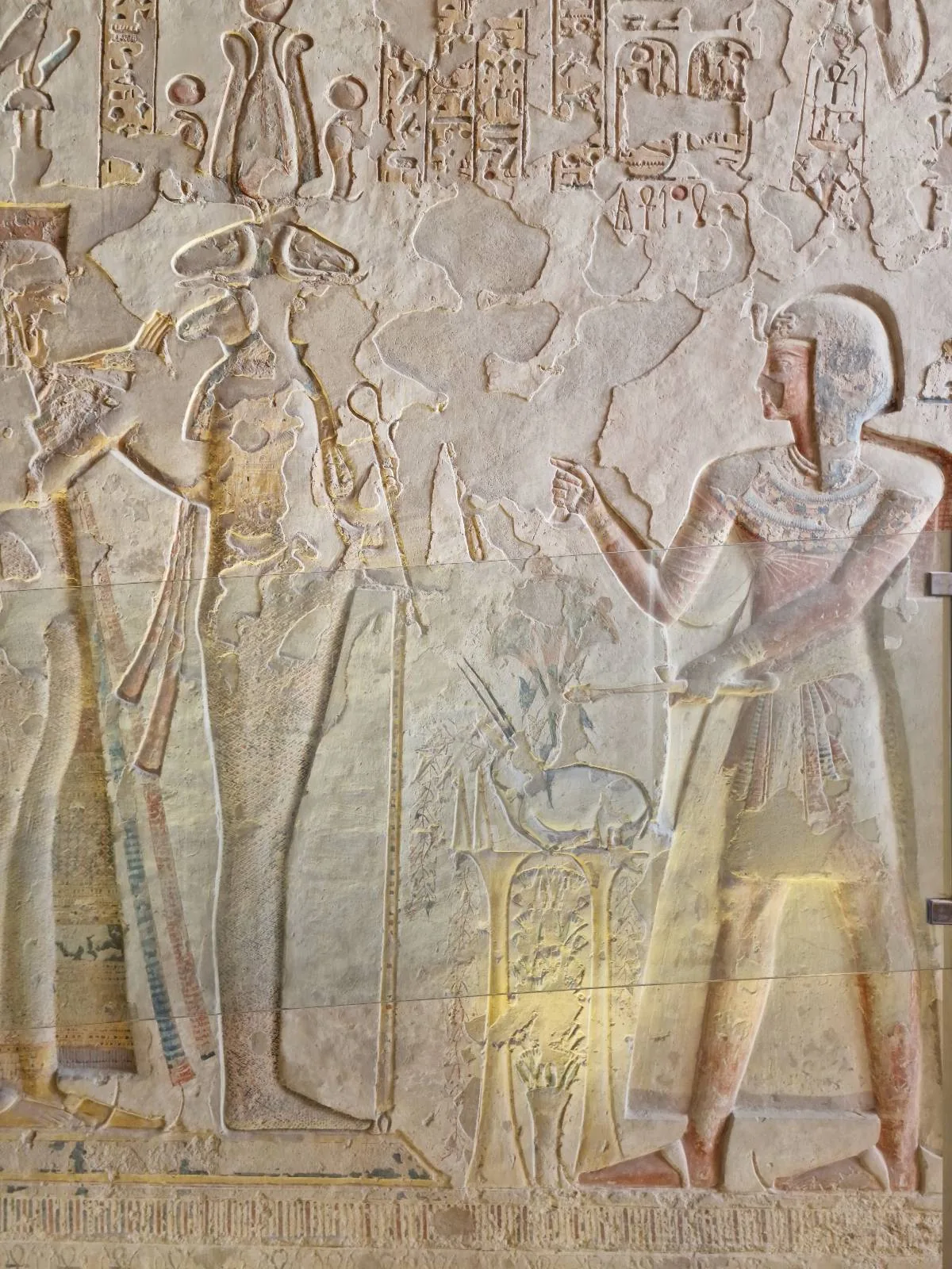 Ancient Egyptian relief depicting two figures standing, facing each other. The left figure wears a tall headdress and ornate attire, while the right figure is dressed simply. Hieroglyphics and detailed carvings fill the background.