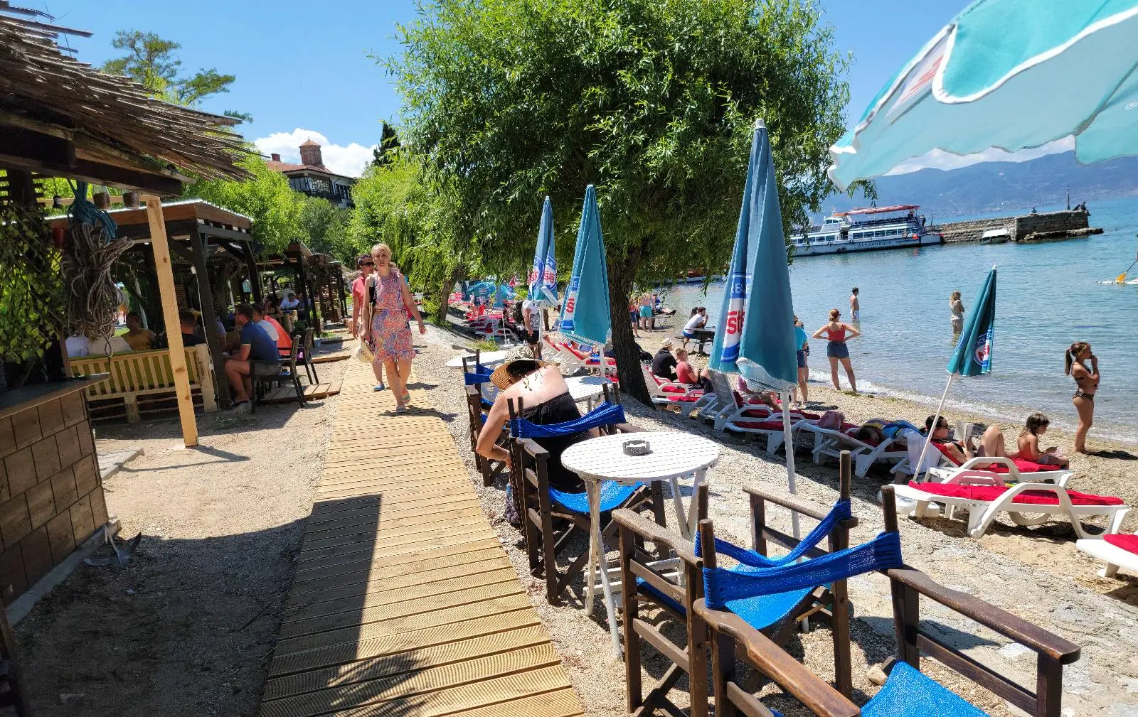 People relax on a sunny beach with sunbeds and umbrellas near the shoreline. Wooden chairs and tables are arranged along a wooden pathway lined with greenery. A boat is visible on the lake in the background under a clear blue sky.