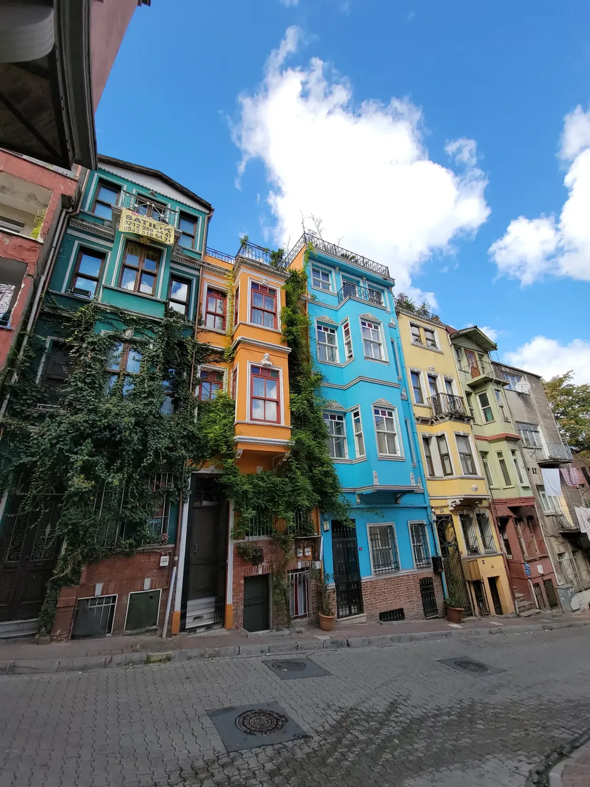 Colorful, narrow houses with ornate facades line a steep street under a bright blue sky with fluffy clouds. Green ivy climbs up one building, adding to the vibrant, lively urban scene.