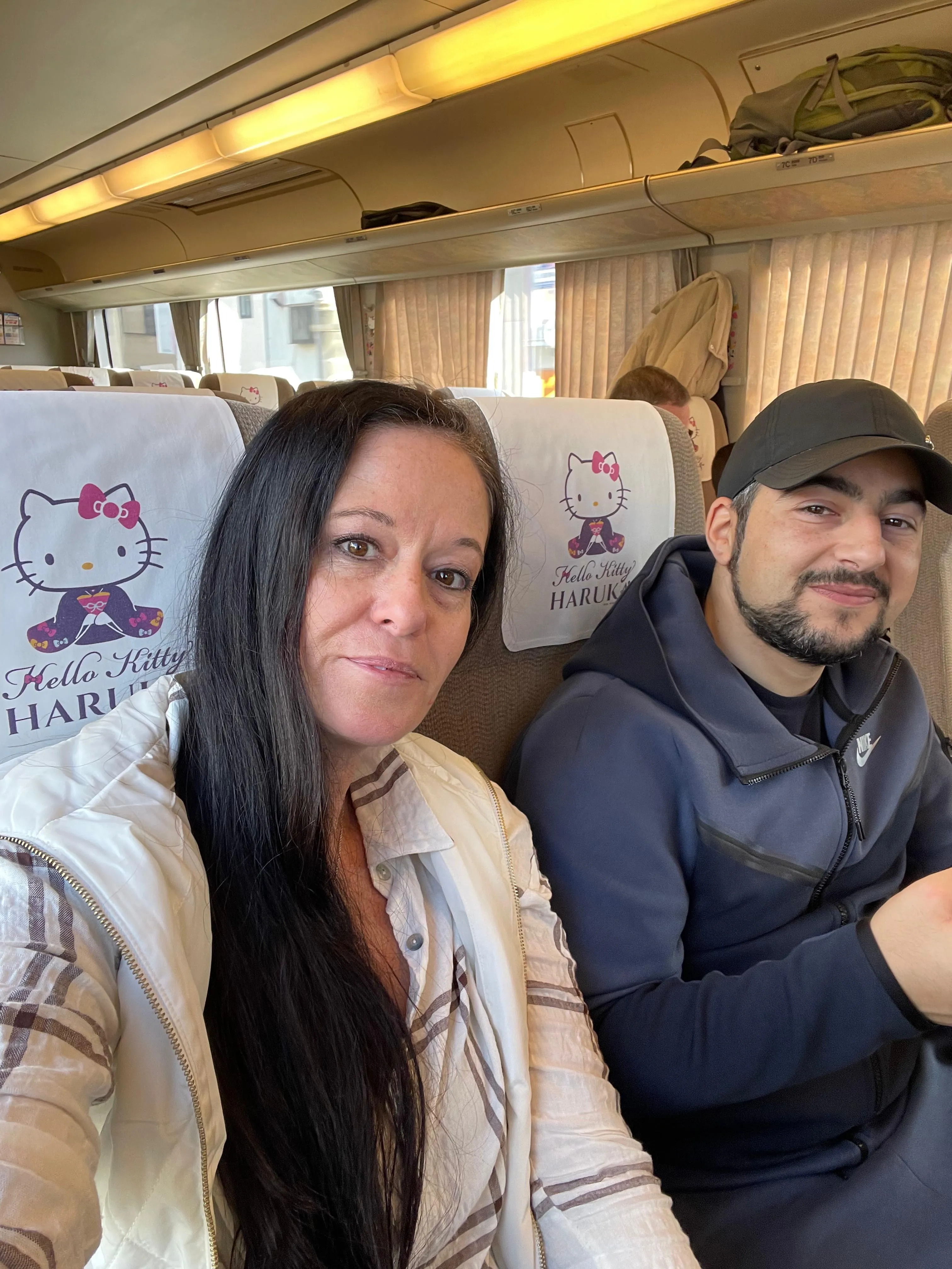 Two people sit side by side on a train with Hello Kitty-themed seat covers. The woman in front has long dark hair and wears a light jacket, while the man beside her wears a dark cap and smiles at the camera.