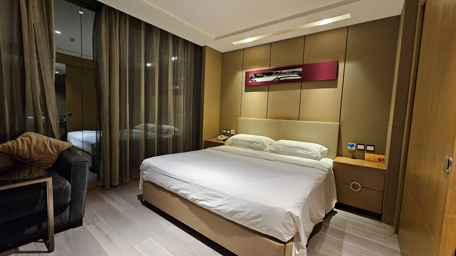 Modern hotel room with a large bed, white linens, two bedside tables, a couch on the left, floor-to-ceiling windows with sheer curtains, and a contemporary wall art piece above the headboard.