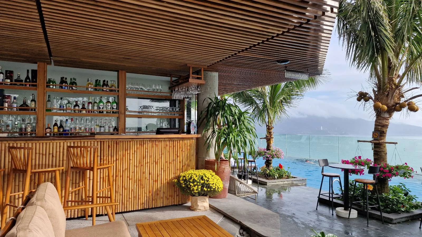 A cozy beachfront bar made of wood overlooks the ocean. It features shelves with bottles, two bar stools, and a potted plant. Palm trees and vibrant flowers frame a clear blue pool and the sea under a cloudy sky.