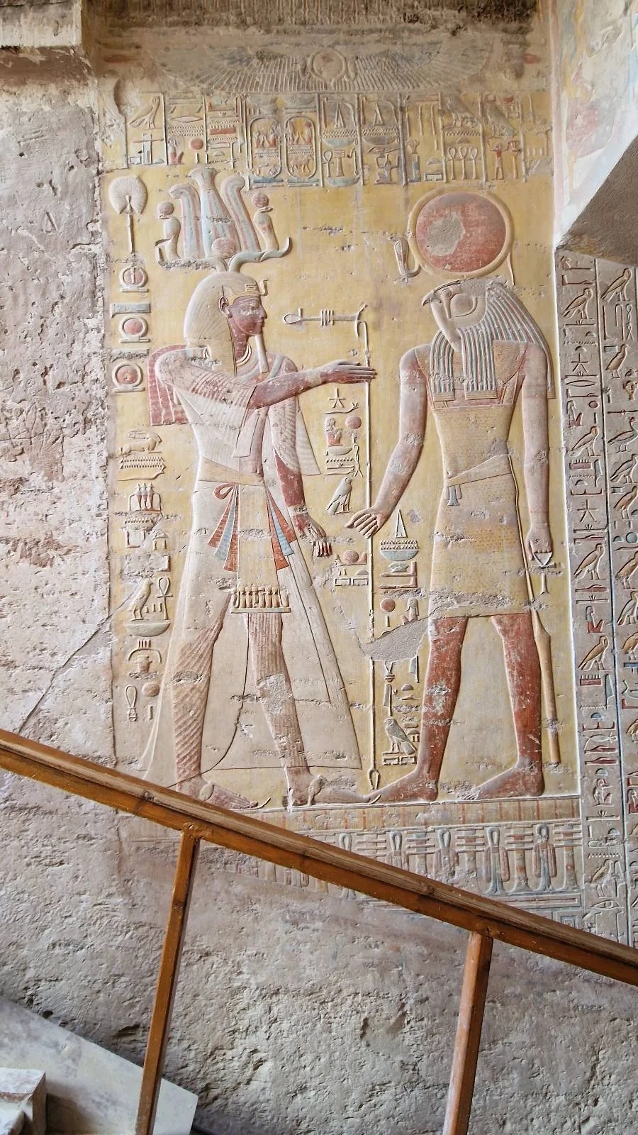 Ancient Egyptian wall art depicting two figures in traditional attire with hieroglyphics above them. The relief shows intricate details in clothing and headpieces against a textured stone background. Wooden railing visible in the foreground.