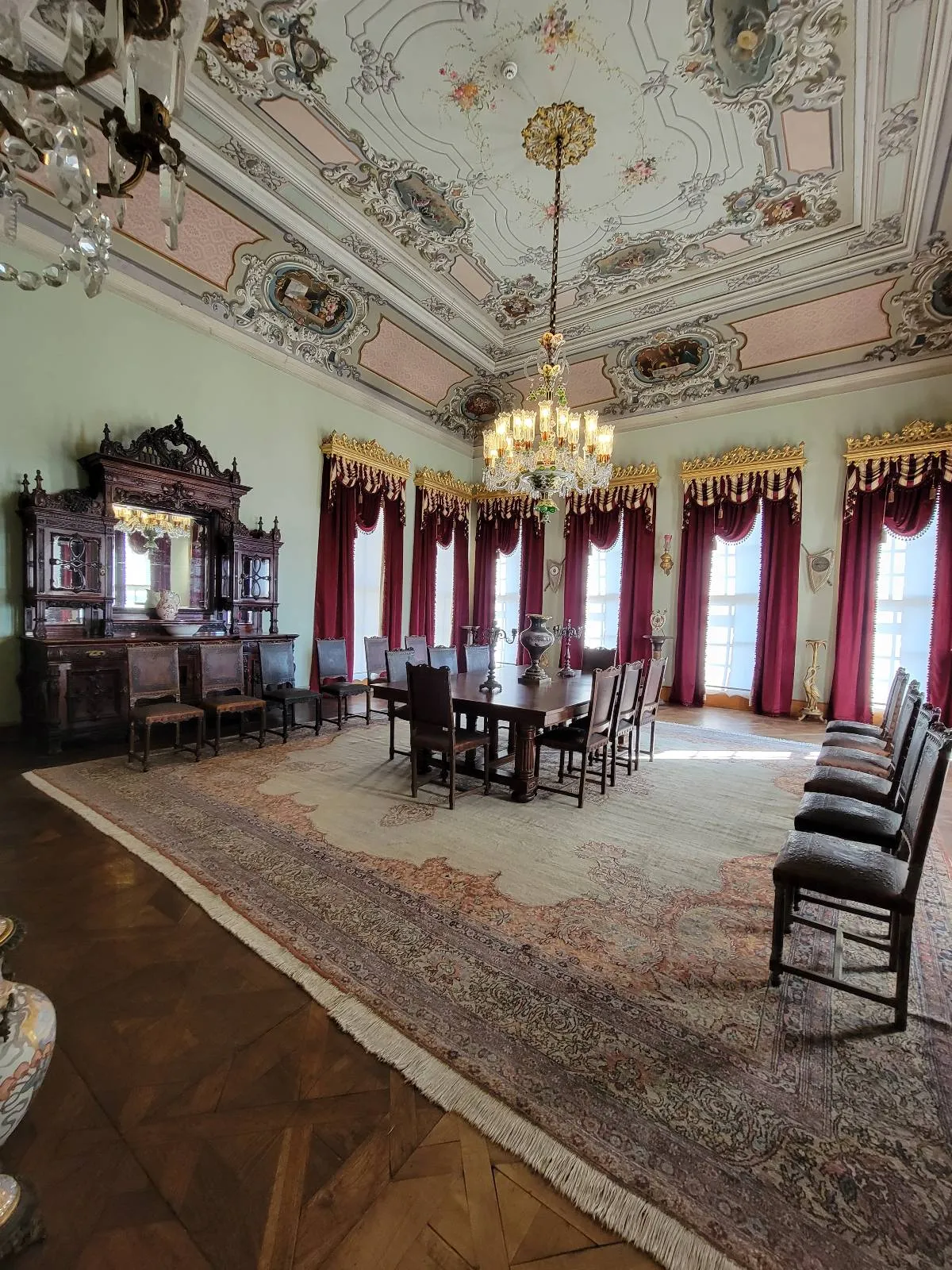A grand dining room with a long table and chairs, ornate ceiling, chandelier, and large windows with red curtains. Vintage furniture and intricate rugs adorn the space.