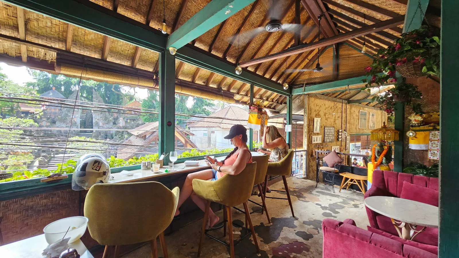 A cozy, open-air café with wooden beams, colorful chairs, and people sitting at tables overlooking a lush, green landscape through large windows.