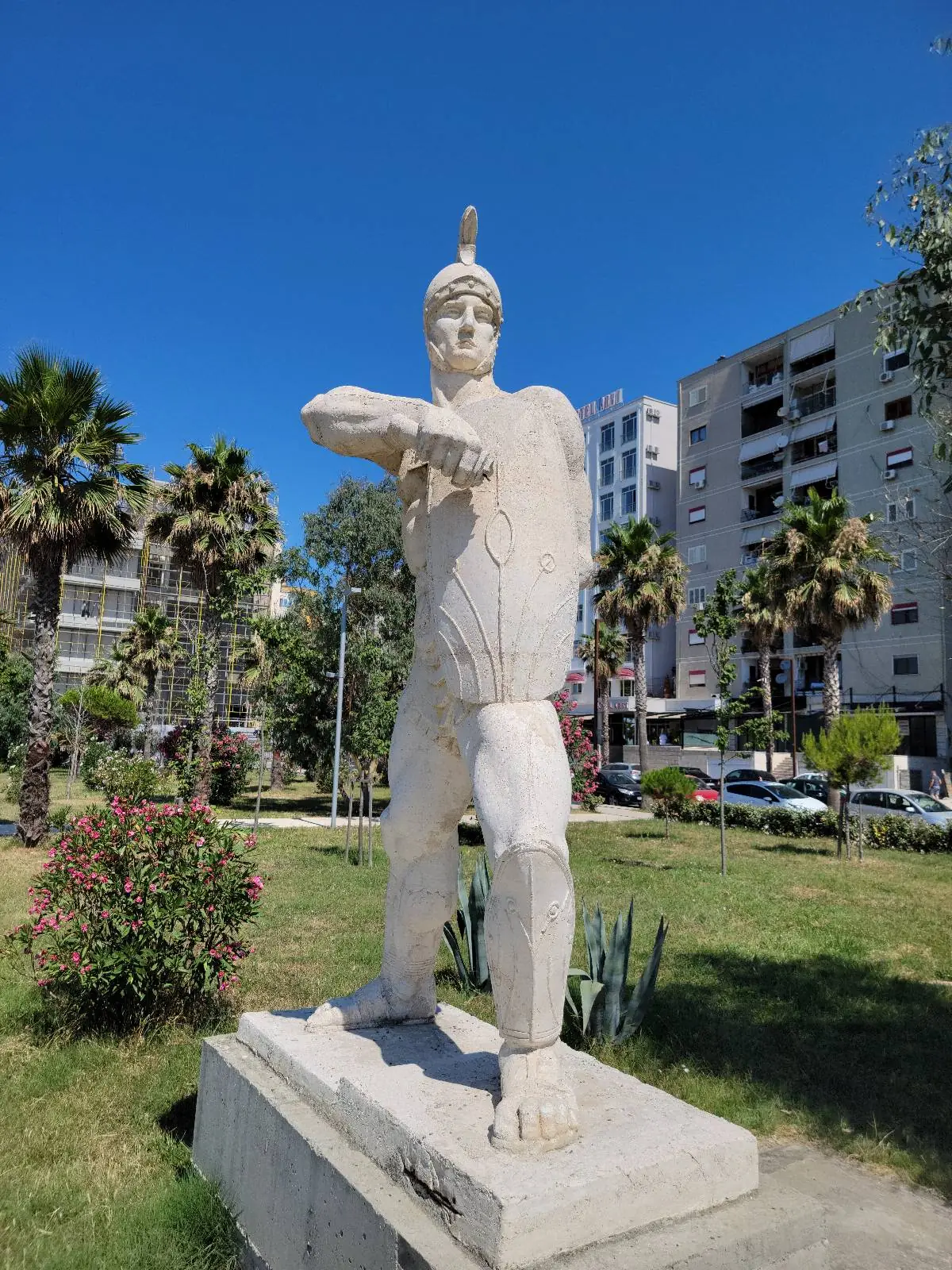 A white statue of a soldier in armor holding a shield stands on a pedestal in a grassy park, with palm trees and modern apartment buildings in the background under a clear blue sky.