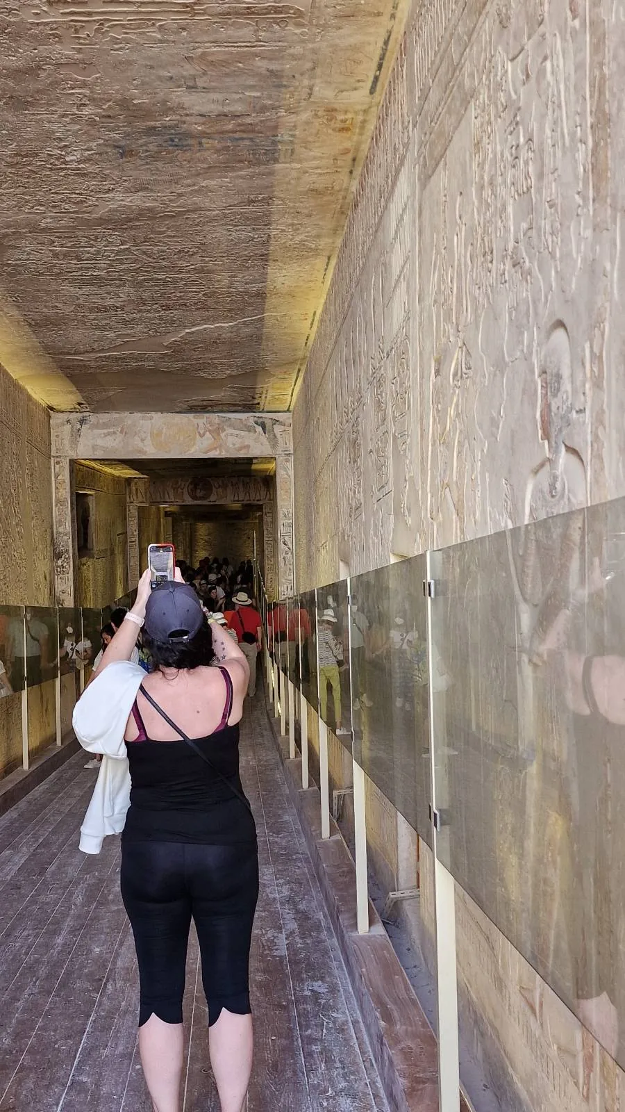 A person wearing black clothing and a hat stands in a narrow hallway taking a photo. The walls are adorned with carvings behind glass panels. The ceiling is ornately decorated, and there is soft lighting throughout the corridor.
